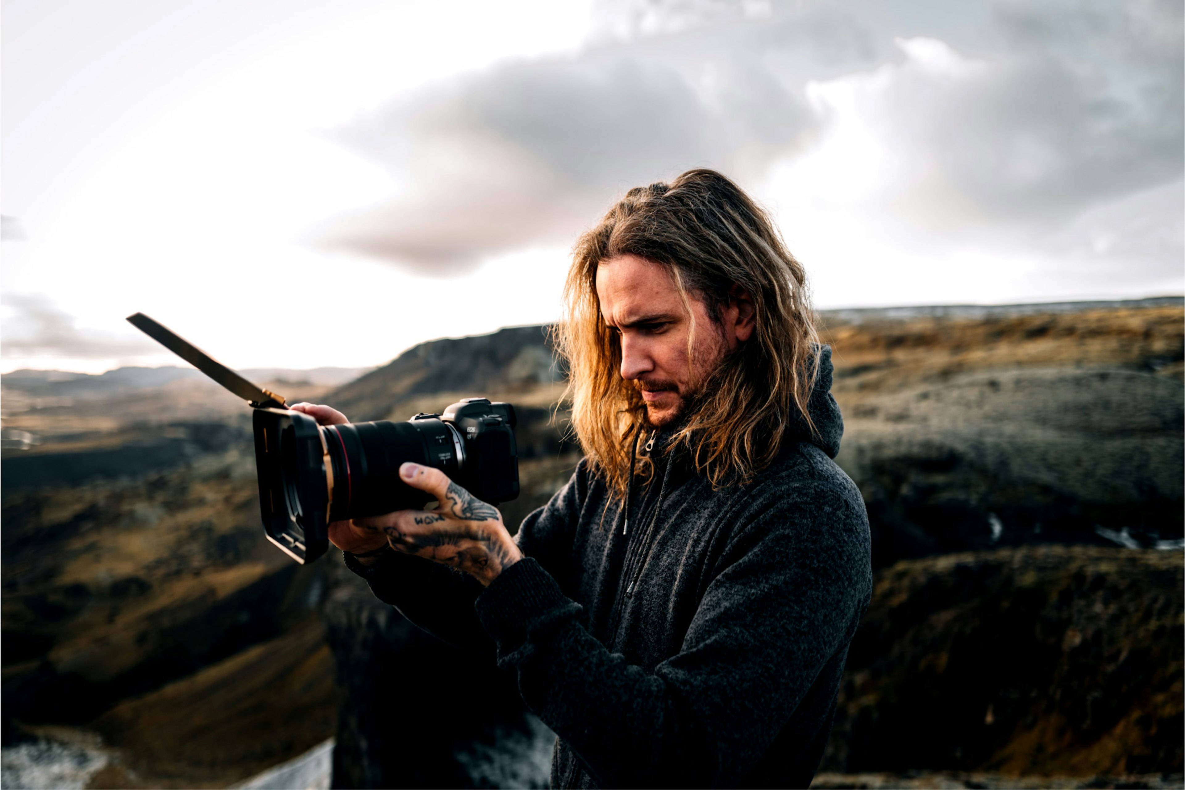 A content creator with long hair focuses on his camera, capturing the scenic beauty of an outdoor landscape, perfect for stock footage